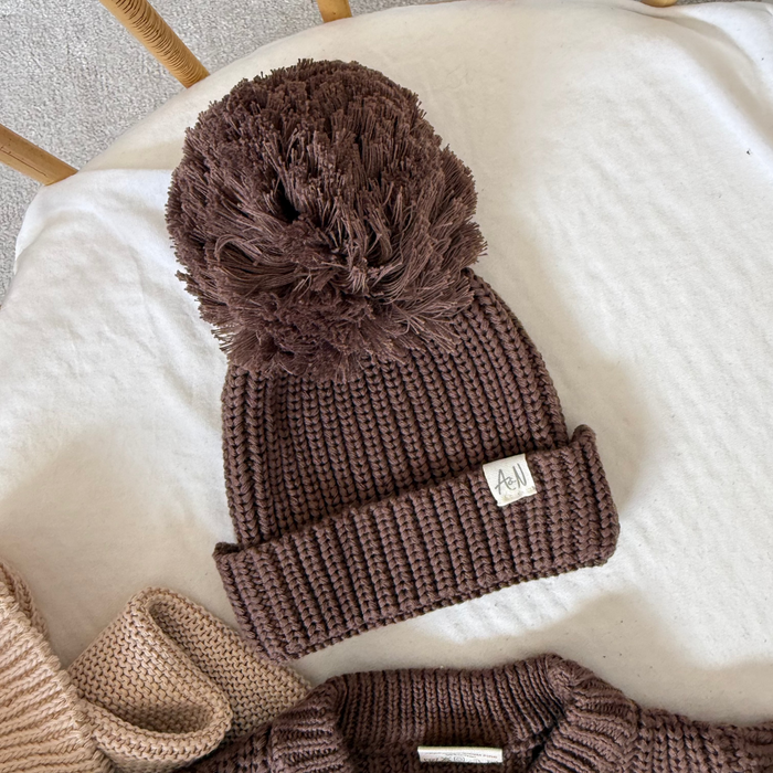 Brown knitted baby beanie with ribbed cuff, side label, and large pom-pom