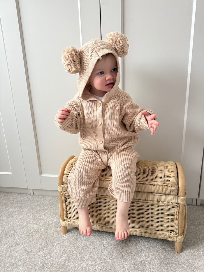 Beige hooded knit romper with oversized pom-pom hood, button front and long legs