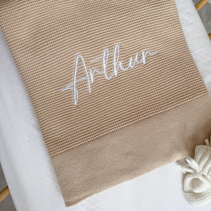 Beige knit baby blanket with white embroidered name 'Arthu' and neat folded edge