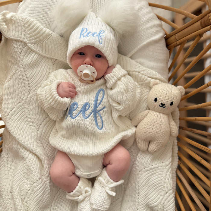 Baby white knitted jumper and hat with script 'Reef', bunny plush beside