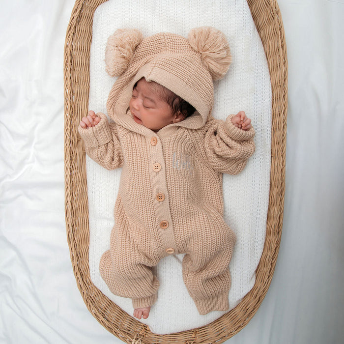 Beige knit romper with button placket and hood, topped with two pom pom beanie