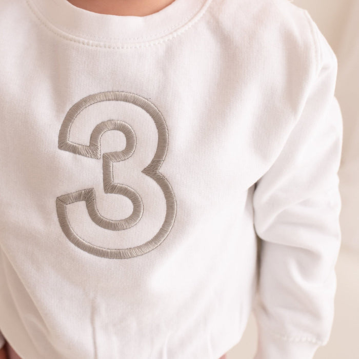 White crewneck sweater close-up with stitched '3' embroidery