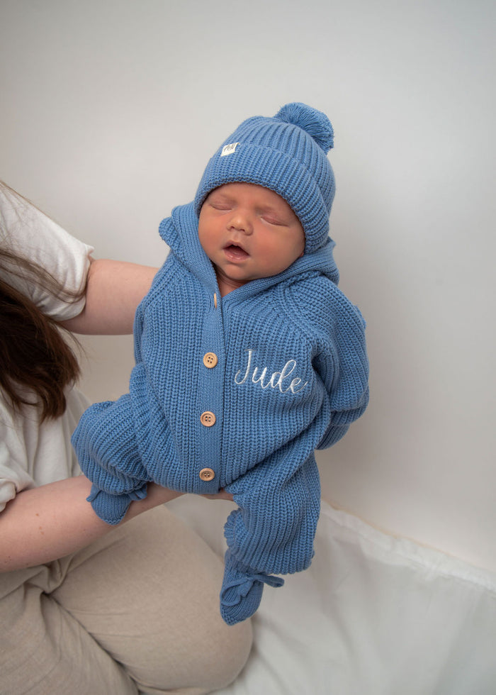 Blue knitted romper with hood and pom-pom beanie, button placket, “Jude” on chest