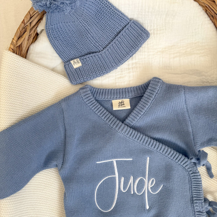 Blue knit romper close-up with 'Jude' script and crossover ties, plus matching hat.