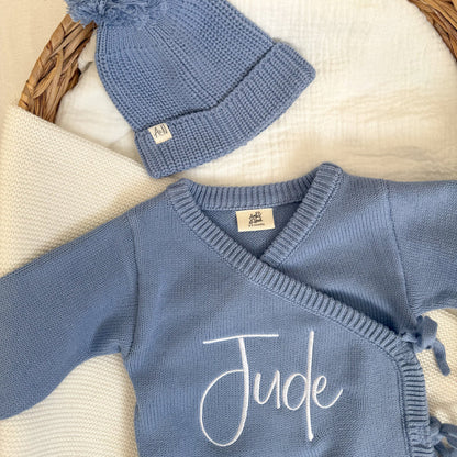 Blue knit romper close-up with 'Jude' script and crossover ties, plus matching hat.