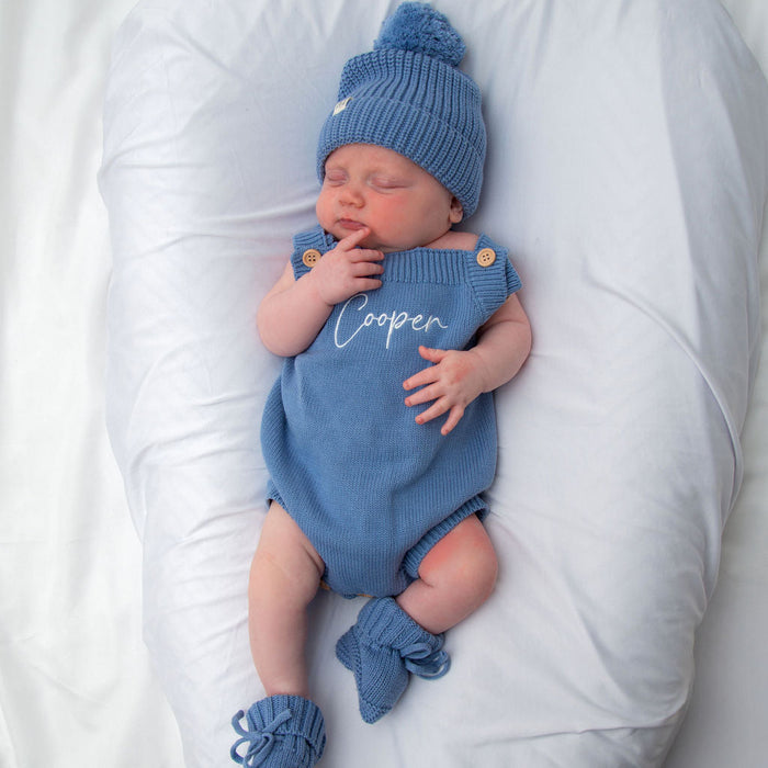 Blue knit personalised romper with button shoulders and matching blue beanie, hand at mouth