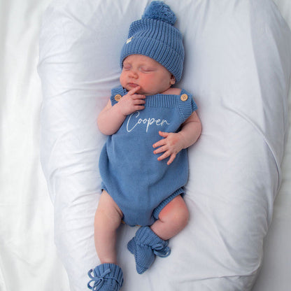 Blue knit personalised romper with button shoulders and matching blue beanie, hand at mouth