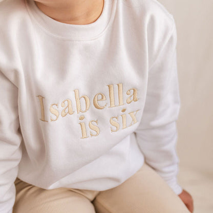 White kids sweater close-up with 'Isabella is six' gold lettering