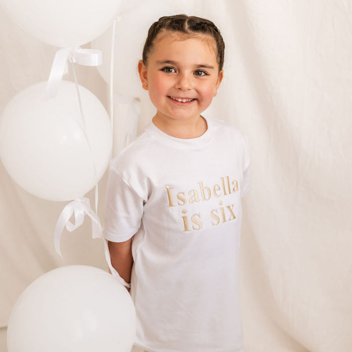Kids white T-shirt with “Isabella is six” chest print, slight side view