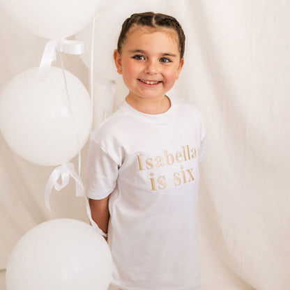 Kids white T-shirt with “Isabella is six” chest print, slight side view