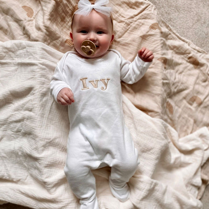White baby romper with “Ivy” embroidered name and long sleeves; baby lying down.