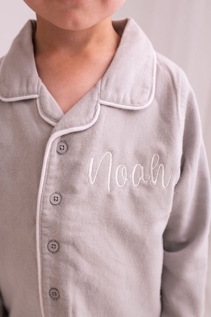 Grey pajama shirt close-up with white trim and cursive 'Noah' embroidery on chest.