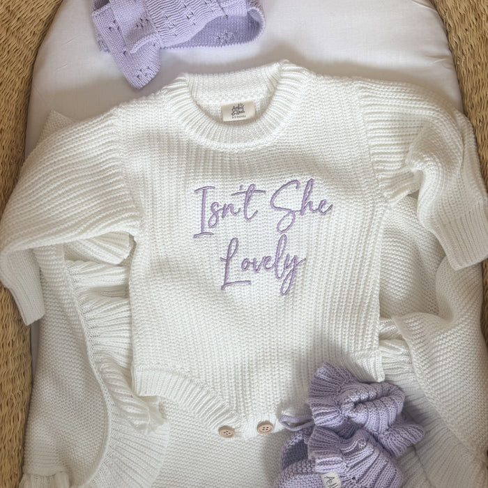 Cream knitted romper front with script 'Isn't She Lovely', two buttons, purple bow