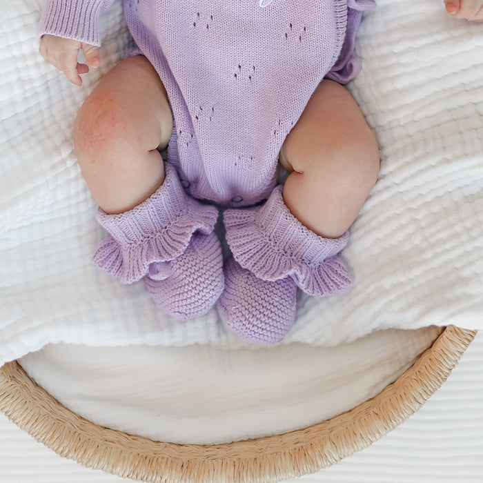 Lilac knitted baby booties with frilled cuffs, worn on infant feet
