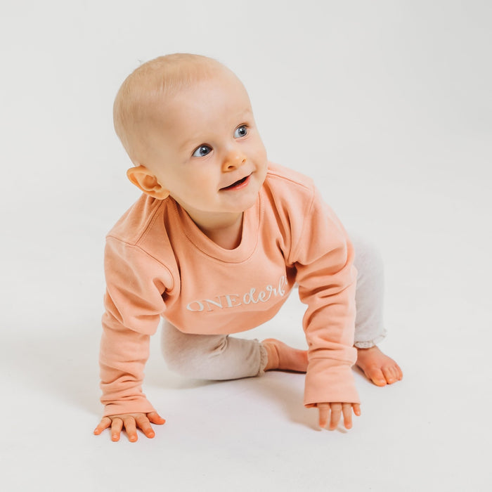 Peach baby birthday sweater with OneDerful front graphic, crawling pose