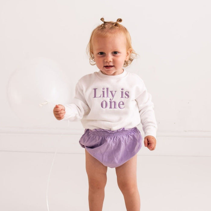 Baby birthday sweater with purple text “Lily is one” on white long-sleeve top