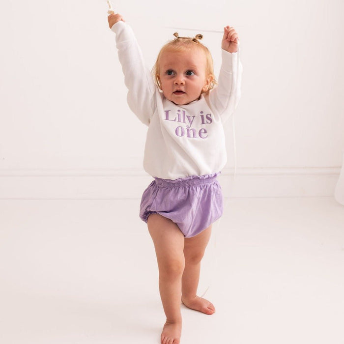 Baby birthday sweater, white long-sleeve with “Lily is one” purple front text