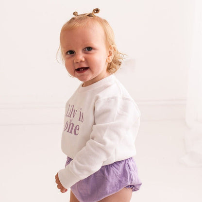 Baby birthday sweater in white with “Lily is one” purple printed design, long sleeves