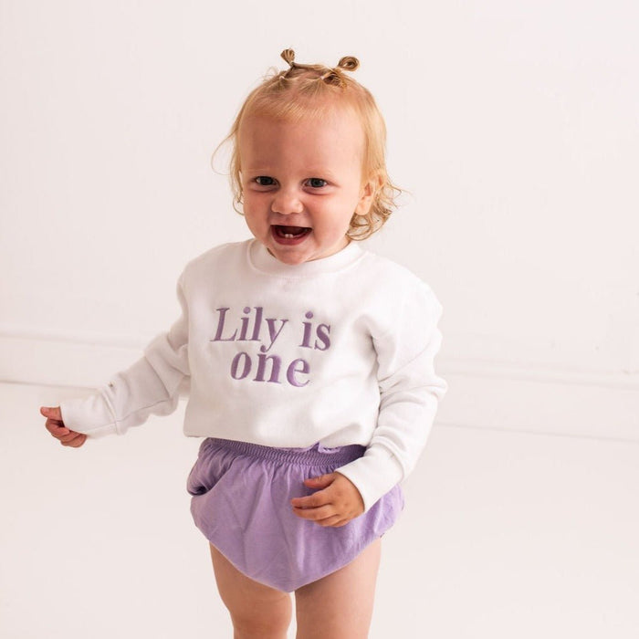 Baby birthday sweater, white long-sleeve with “Lily is one” purple text at chest