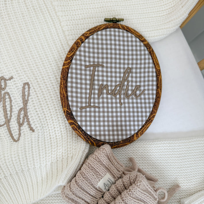 Oval baby name announcement hoop with brown trim and embroidered “Hello” on gray gingham
