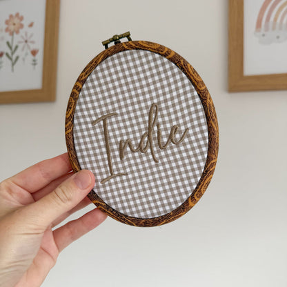 Oval baby name announcement hoop with brown trim and embroidered “Hello” on gray check cloth