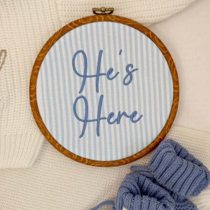 Round baby name sign with blue striped background, ‘Here’s Here’ cursive, brown rim