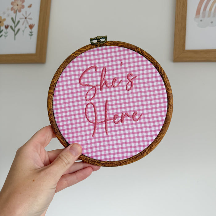 Round pink gingham baby announcement sign with stitched text: “She's Here”.