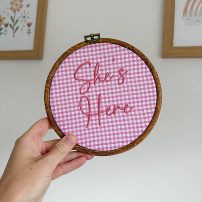 Round pink gingham baby announcement sign with stitched text: “She's Here”.