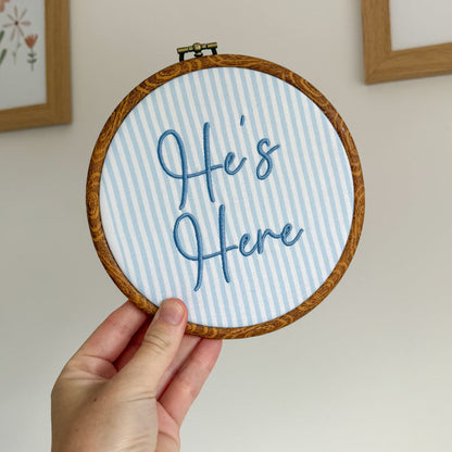 Round baby name sign, blue striped insert reading ‘Here’s Here’ in cursive, held in hand