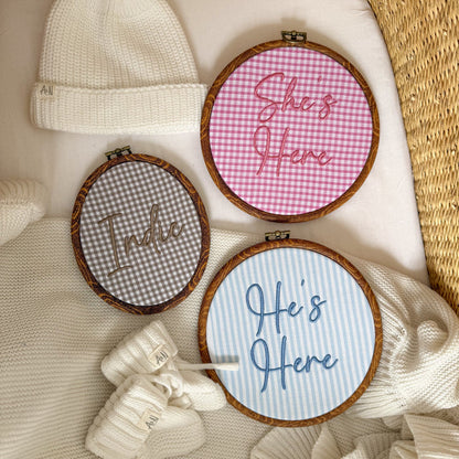 Three round baby name signs: pink gingham ‘She’s Here’, blue ‘Here’s Here’, gray name