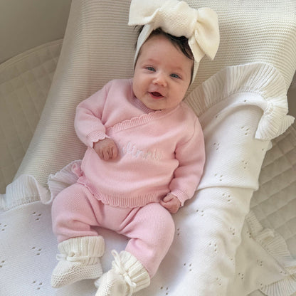 Baby in pink knit kimono set with ruffled neckline, sitting on cream quilted cushion