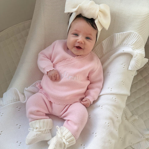 Baby in pink knit kimono set with ruffled neckline, sitting on cream quilted cushion