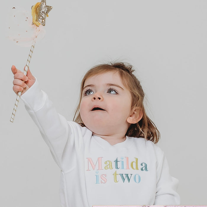 White personalised birthday sweater with 'Matilda is two' rainbow-style text print