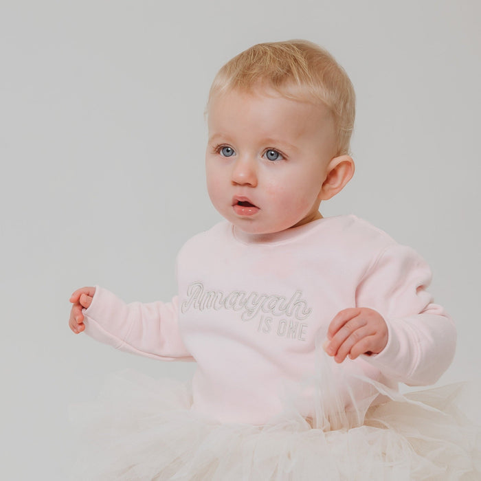 Light pink baby sweatshirt with glittery name and 'is one' print