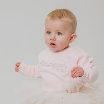 Light pink baby sweatshirt with glittery name and 'is one' print