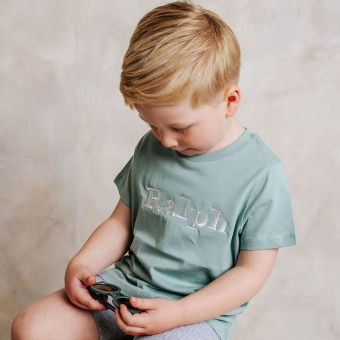 Sage green personalized T-shirt with 'Ralph' front print, child seated looking down