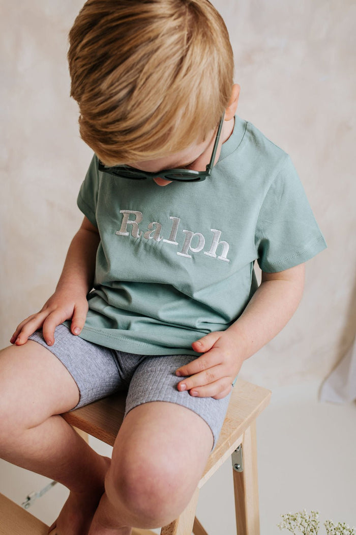 Sage green personalized T-shirt with 'Ralph' back print, child seated on chair