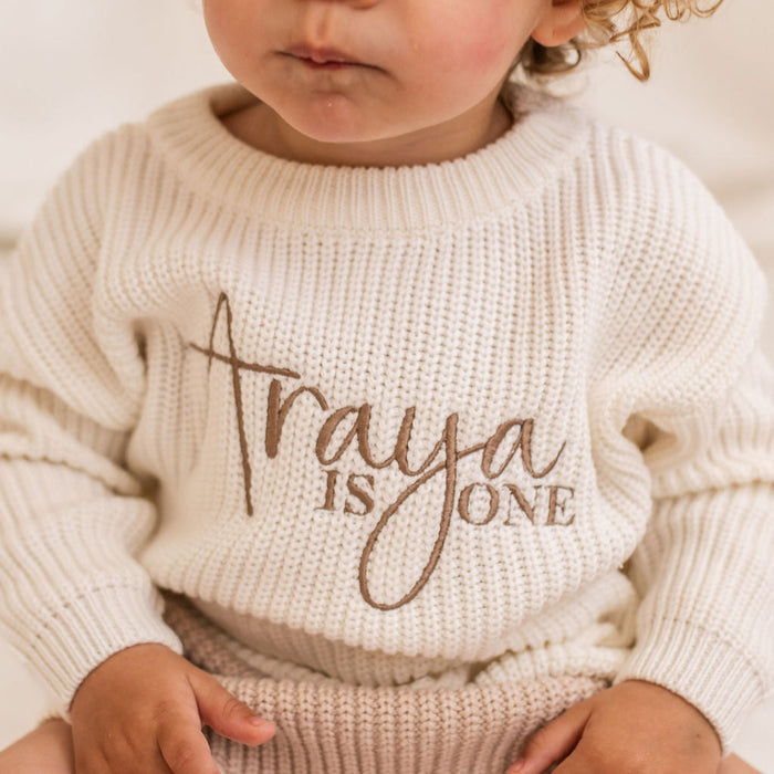 White knitted baby jumper with ribbed crew neck and script 'is ONE' chest print