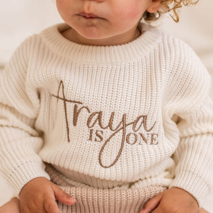 White knitted baby jumper with ribbed crew neck and script 'is ONE' chest print