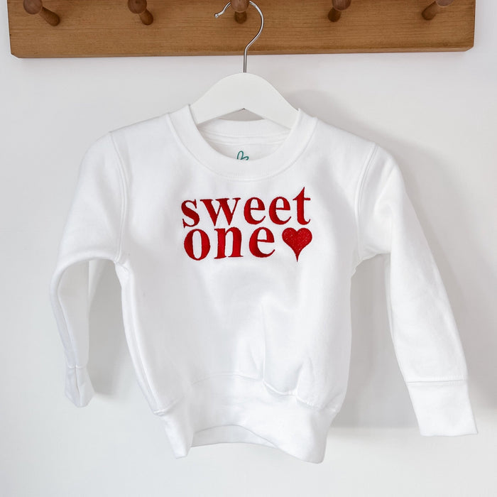 White crewneck sweater with 'sweet one' red text and heart motif at chest
