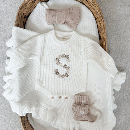 White knitted baby romper with embroidered floral letter, ruffled hem, and bow detail