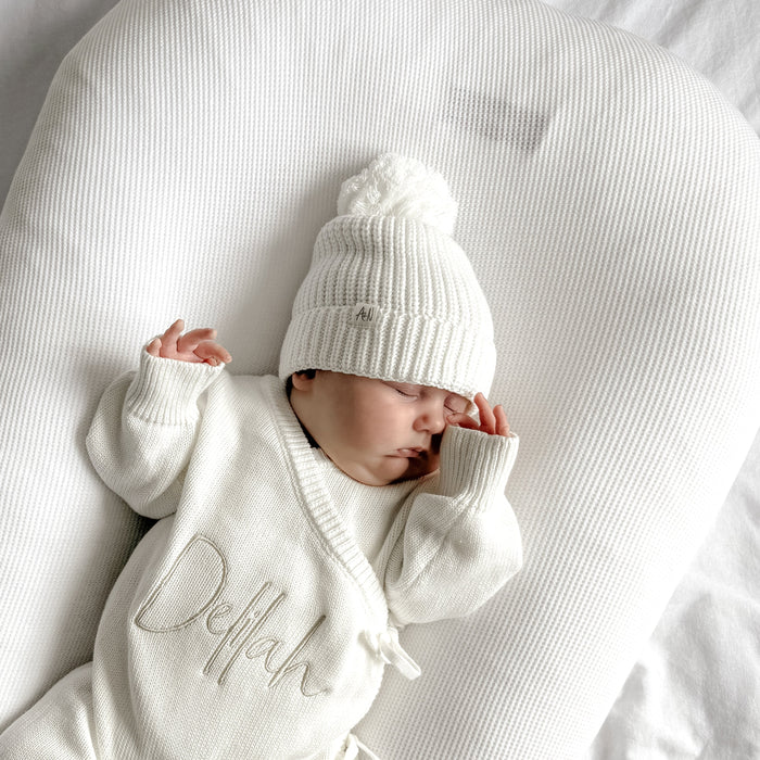 White knitted baby beanie with ribbed brim and pom-pom, worn on baby’s head