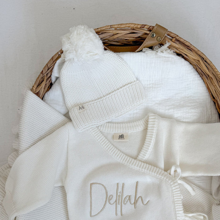 White knitted baby hat with ribbed brim and white pom-pom in basket