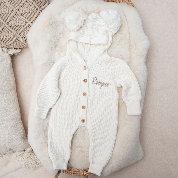 White knitted baby romper with hood, faux-fur trim, pom detail, and chest name embroidery