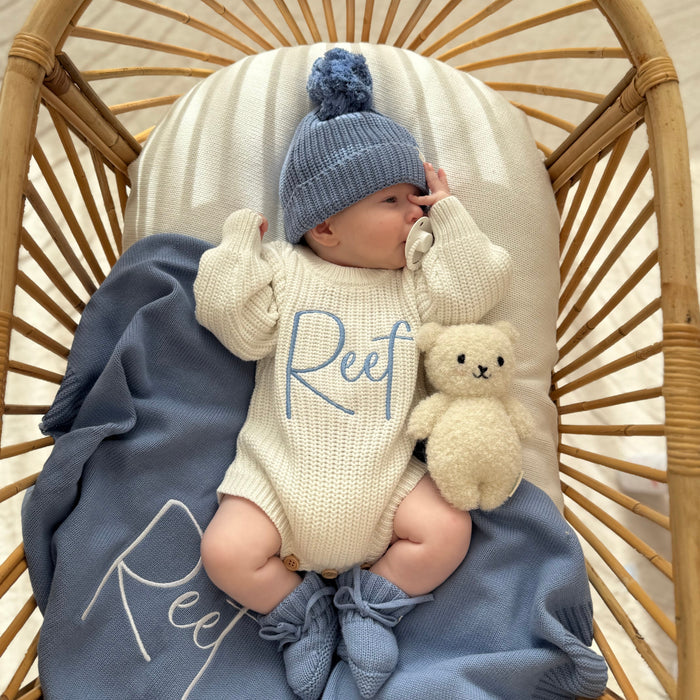 Cream knitted romper with blue 'Reef' front, blue knit hat, and plush rabbit toy