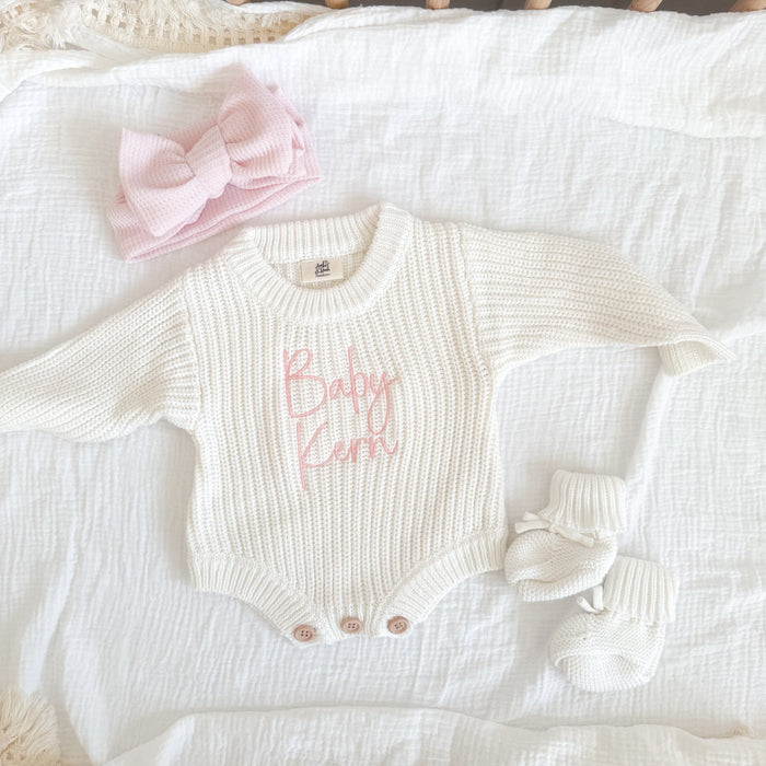 White knitted baby romper with pink "Baby" text, bow accents and snap buttons